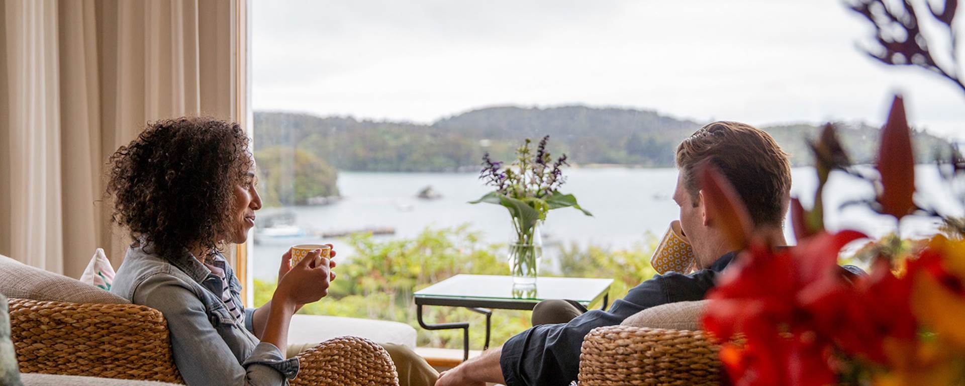 A couple sit by the window of the Stewart Island lodge enjoying a cup of tea and looking at the view