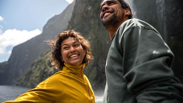 A couple smile while taking in the view of Milford Sound, with a waterfall cascading behind them